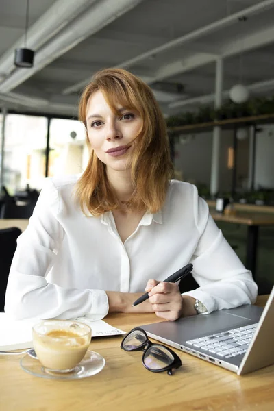 Masada oturan beyaz tişörtlü, kırmızı saçlı genç bir kadın elinde siyah bir kalem ve masada bir defterle.