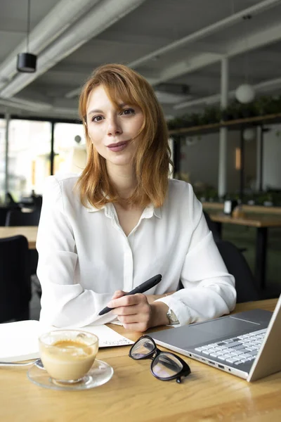 Masada oturan beyaz tişörtlü, kırmızı saçlı genç bir kadın elinde siyah bir kalem ve masada bir defterle.