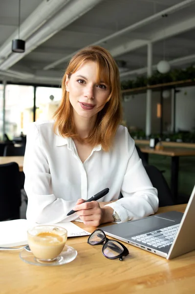 Kızıl saçlı güzel kadın ahşap masada oturuyor ve fonda kafesi olan şeffaf bardaktan kahve içiyor.