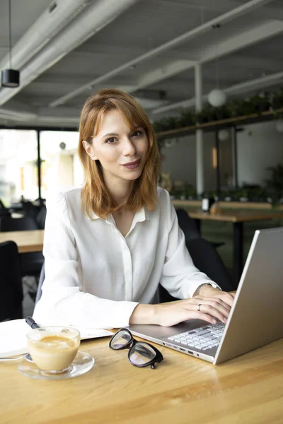 Gözlüklü güzel bir iş kadını masada dizüstü bilgisayarla oturuyor.