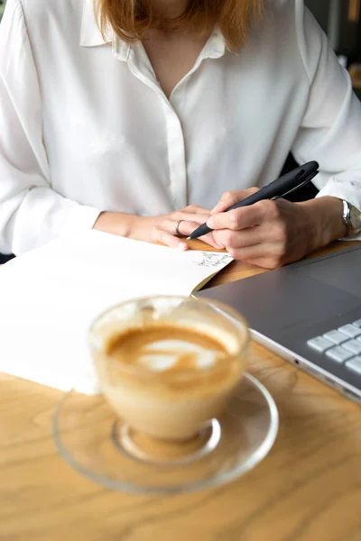 Güzel bir iş kadını ön planda bir fincan kahveyle kalemle not alıyor.