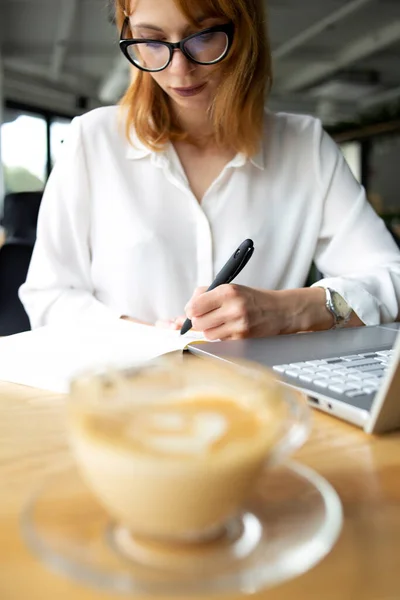 Güzel bir iş kadını ön plandaki kahve ve dizüstü bilgisayara not alıyor.