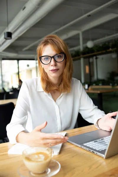 Gözlüklü ve beyaz gömlekli genç bir kadın halka açık bir yerde oturuyor.