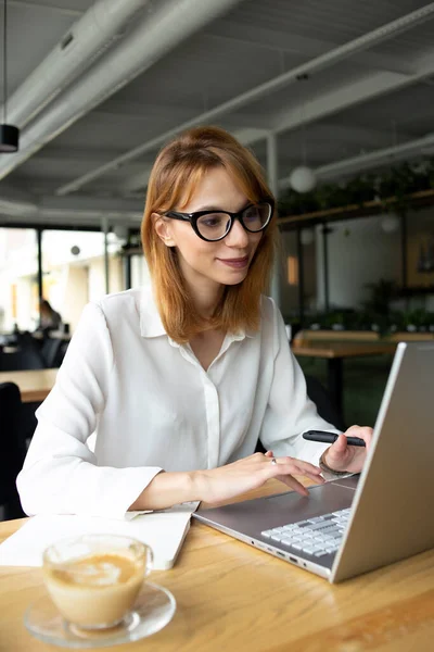 Masada oturan, beyaz tişörtlü, beyaz tenli, gri dizüstü bilgisayarı deviren, masada bir fincan kahve olan beyaz, zayıf bir kadın.