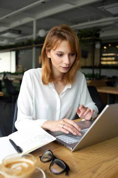 Masada oturan, beyaz tişörtlü, beyaz tenli, gri dizüstü bilgisayarı deviren, masada bir fincan kahve olan beyaz, zayıf bir kadın.