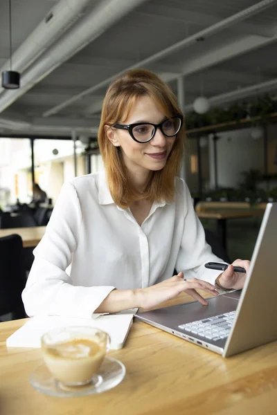 Gözlüklü ve beyaz gömlekli genç bir kadın halka açık bir yerde oturuyor.