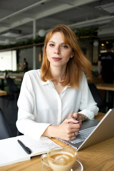 Beyaz gömlekli genç iş kadını masada oturmuş elinde saatlerle kahve kapuçinosu içiyor.