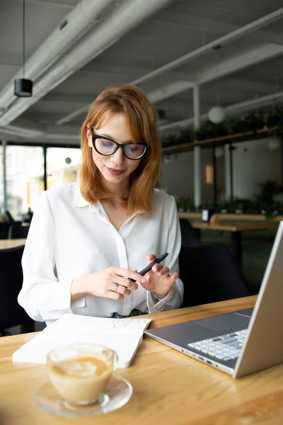 Gözlüklü ve beyaz gömlekli genç bir kadın halka açık bir yerde oturuyor.