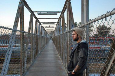 Siyah ceketli, uzun saçlı ve eşarplı genç adam köprüde.