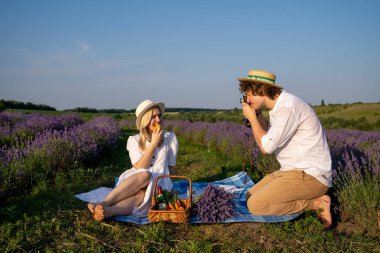 two persons couple in matching outfit have picnic in lavender field. They have fun, laugh and are feeding each other for photo session. Romantic relationship concept