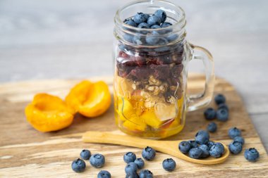 blur background, healthy organic breakfast, transparent glass cup presentation, with mix of cut fruits and nuts: blueberries, berries, walnuts, apricot, peach, cranberry  on a wooden plate