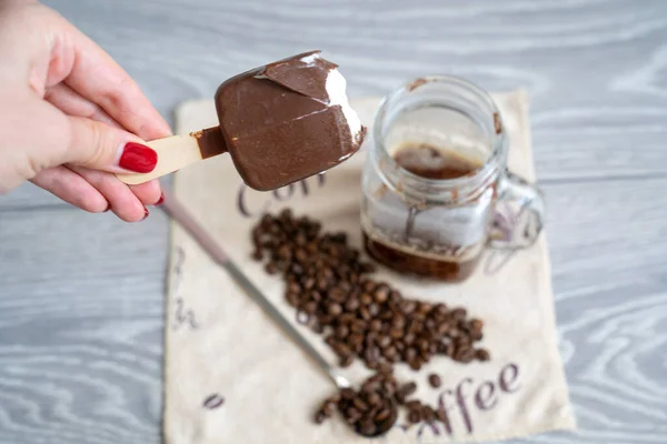 blur background, woman hand with manicure holds half eaten ice cream and pours it into strong black coffee espresso shot in glass cup, coffee set on table, long spoon and coffee beans on napkin, summer refreshment affogato