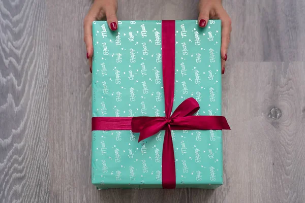 woman hand holding green turquoise color gift box with red ribbon bow on grey wooden background. Concept: holiday, celebration, birthday, New Year, Christmas, Valentines Day, Mothers Day, Thanksgiving Day