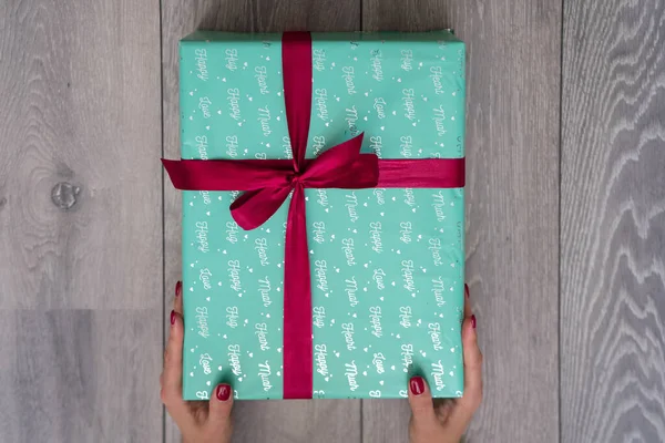 woman hand holding green turquoise color gift box with red ribbon bow on grey wooden background. Concept: holiday, celebration, birthday, New Year, Christmas, Valentines Day, Mothers Day, Thanksgiving Day