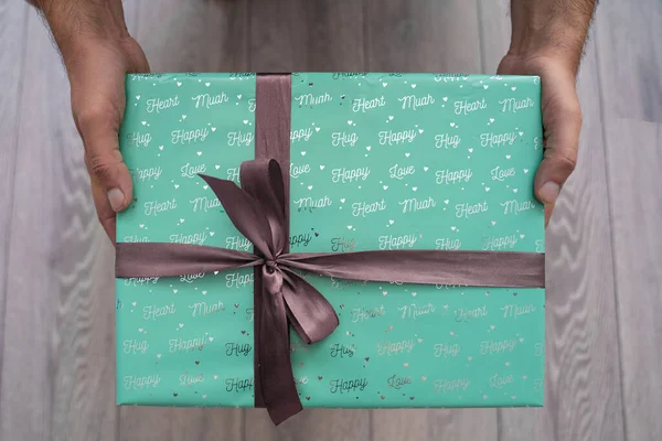 man hand holding green turquoise color gift box with grey ribbon bow on grey wooden background. Concept: holiday, celebration, birthday, New Year, Christmas, Valentines Day, Mothers Day, Thanksgiving