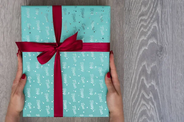 woman hand holding green turquoise color gift box with red ribbon bow on grey wooden background. Concept: holiday, celebration, birthday, New Year, Christmas, Valentines Day, Mothers Day, Thanksgiving