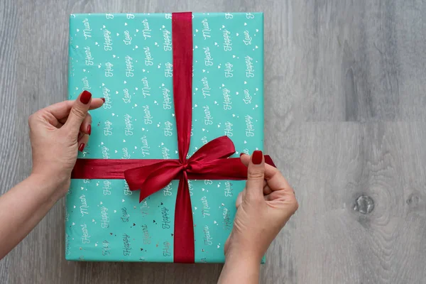 woman hand holding green turquoise color gift box with red ribbon bow on grey wooden background. Concept: holiday, celebration, birthday, New Year, Christmas, Valentines Day, Mothers Day, Thanksgiving