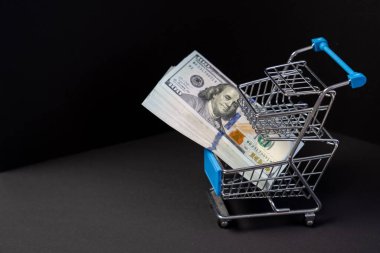 Paralı alışveriş arabası, siyah arka planda yüz dolarlık banknotlar, satın alma konsepti, para harcaması, kara Cuma indirimi, para birimi