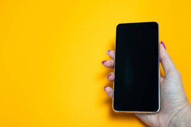 Tırnak manikürlü bir kadın elinde akıllı telefon, sarı arka planda boş siyah ekran, mesaj için yer, resim, modern teknoloji, internet bağlantısı, wi-fi, mesajlaşma 