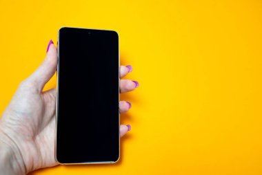 Tırnak manikürlü bir kadın elinde akıllı telefon, sarı arka planda boş siyah ekran, mesaj için yer, resim, modern teknoloji, internet bağlantısı, wi-fi, mesajlaşma 