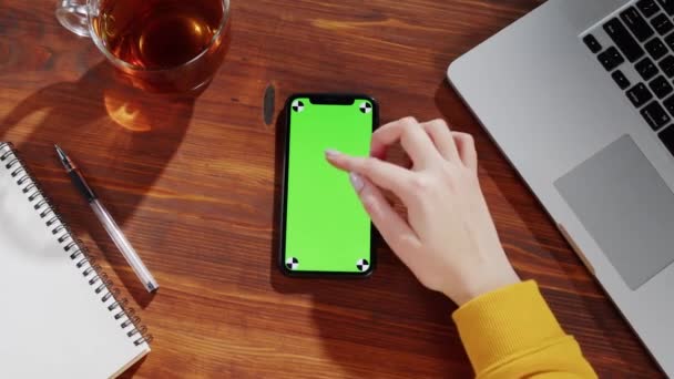 女性の手は携帯電話 緑の画面 クロマキー メモ帳 ノートパソコンをテーブルの上に押します — ストック動画