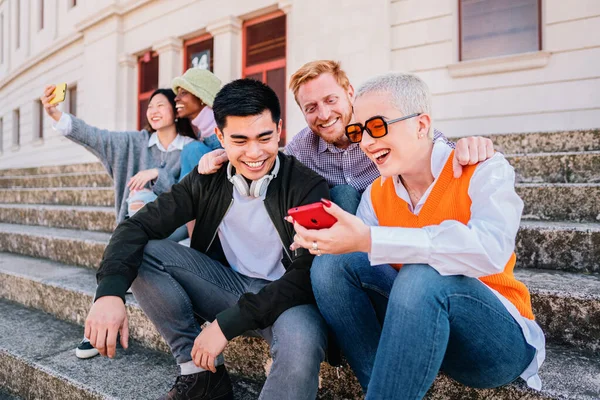 Ungdomar som sitter utomhus och tittar på sociala medier i smarttelefonen. Glada multietniska hipsters ser roligt online-innehåll på mobiltelefon — Stockfoto