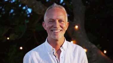 Close up ortrait of happy mature man with gray hair looking and smiling at camera outdoor at sunset in a backyard. High quality 4k slowmotion footage