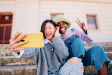 Two joyful cheerful girls taking a selfie while sitting together outdoors and showing peace gesture. high quality photo