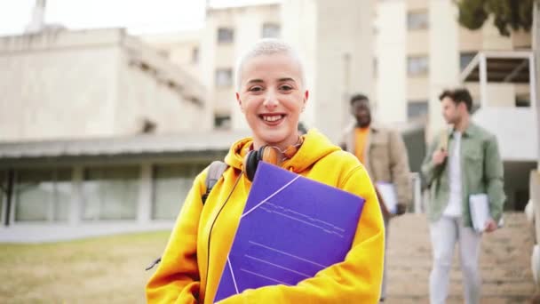 一位留着短发的女学生笑着站在学院前 看着镜头 背后是一群在大学校园里打墙的年轻人 教育概念 是的高 — 图库视频影像