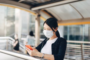 Genç yetişkin iş kadını Asyalı kadın virüs koronasını veya covid19 'u ve hava kirliliğini korumak için yüz maskesi takıyor. Cep telefonu uygulaması kullanılıyor. Gündüz konseptinde halk sağlığı ile kentsel yaşam tarzı.