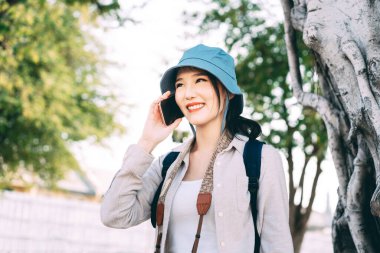 Çekici mutlu gülümseme genç Asyalı kadın gezgin mavi şapka ve sırt çantası takıyor. Aramak için cep telefonu internetini kullanmak. Gündüzleri şehir yaşamında gezen insanlar. 