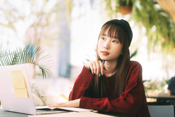 Asian teenager college student woman wear red sweater and wool hat. Using laptop study and work online at cafe. University people city lifestyle at outside from home on day concept.