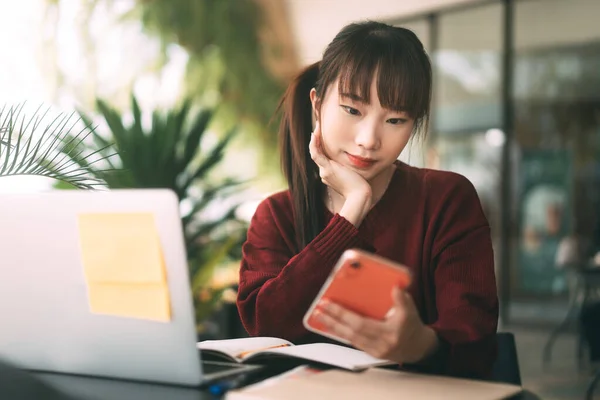 年轻的亚裔女学生穿着红色毛衣 使用手机和笔记本电脑在咖啡店上网学习 大学生日常生活习惯的概念 — 图库照片