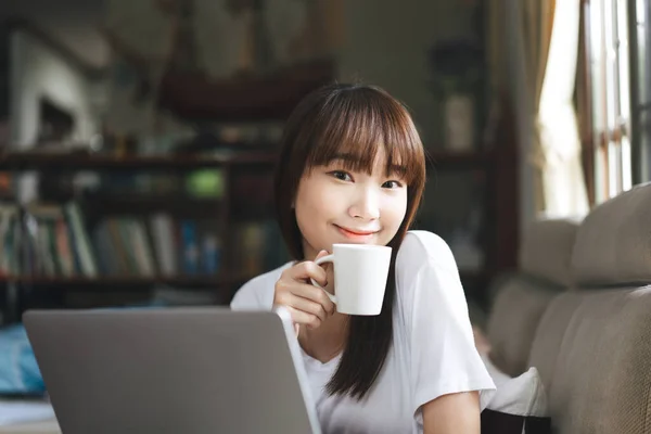 网上学习的学生呆在家里是为了社交上的疏远 快乐的笑着 亚洲少女通过互联网和笔记本电脑一起工作 手里拿着咖啡杯 带窗户灯的客厅背景 — 图库照片