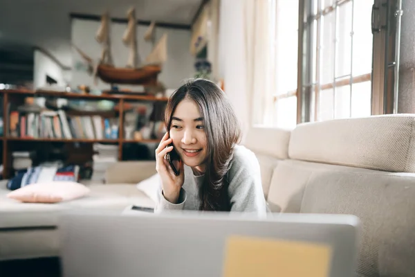 年轻的亚洲自由职业者女商人在家里通过互联网在网上工作 用手机与客户通话 带窗户灯的客厅工作空间背景 — 图库照片