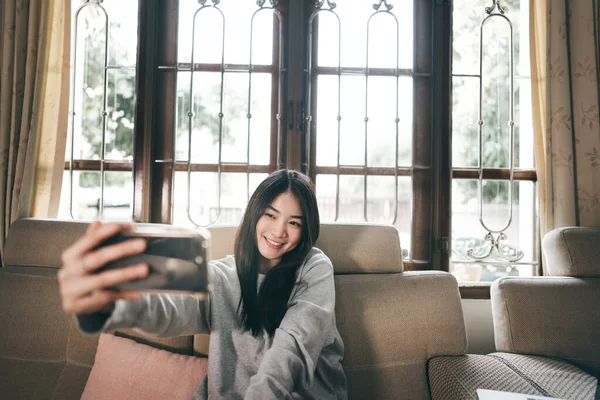 幸せな笑顔若いですアジアの女性自画撮り写真によって携帯電話によって自宅でリビングルームで窓の背景 — ストック写真