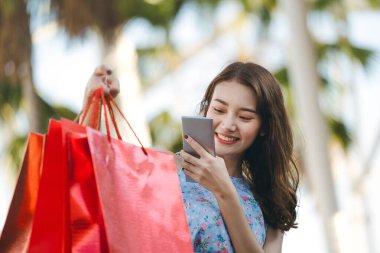 Genç Asyalı etkili kadın açık hava parkından alışveriş yapıyor. Akıllı telefonu kullanarak fotoğrafçı çantasını kullanarak sosyal ağı yüklemek. Yaz indirimi konsepti. Kopyalama alanı olan arkaplan.