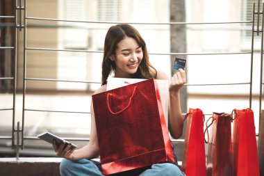 Güzel bir gülümseme genç Asyalı kadın akıllı telefon alışverişi yapar ve internetten kredi kartıyla öder. Bayram indirimi konsepti alışverişi. Şehir kızı yaşam tarzı mutlu açık hava aktiviteleri.