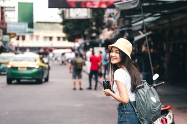 Sırt çantalı, beyaz gömlekli, mutlu Asyalı genç bir kadın. Kamerayı tutuyorum. Khaosan Yolu 'ndaki tarihi cadde pazarının yanında. Bangkok, Tayland 'da şehir kızı yaşam tarzının tadını çıkar.