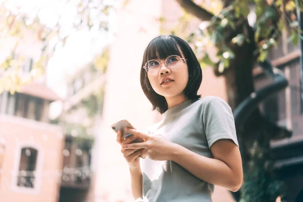 年轻快乐的亚洲女人带着眼镜 用手机进行社交媒体应用 每日在室外发短信 建筑物的背景 — 图库照片