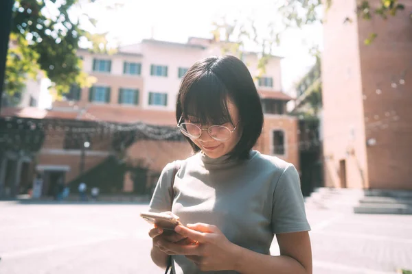 年轻快乐的亚洲女人带着眼镜 用手机进行社交媒体应用 每日在室外发短信 建筑物的背景 — 图库照片