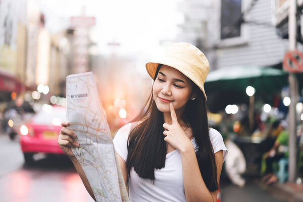 Young adult asian pretty woman solo tourist using map for find route trip. Outdoor travel on day in the city. Background with traffic and bokeh. Bangkok, Thailand.