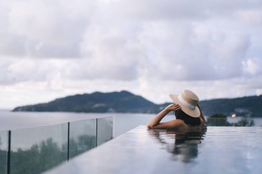 Tatil konseptiyle yaz seyahati. Asyalı kadının arka görüntüsü açık havuz tatil köyü deniz manzarasında rahatlıyor. Hasır şapka ve bikiniyle. Phuket Adası, Tayland Yerli Turizm