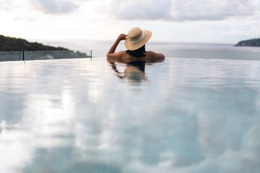 Tatil konseptiyle yaz seyahati. Asyalı kadının arka görüntüsü açık havuz tatil köyü deniz manzarasında rahatlıyor. Hasır şapka ve bikiniyle. Phuket Adası, Tayland Yerli Turizm