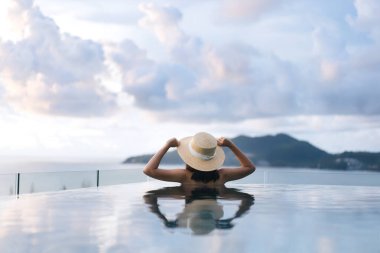 Tatil konseptiyle yaz seyahati. Asyalı kadının arka görüntüsü açık havuz tatil köyü deniz manzarasında rahatlıyor. Hasır şapka ve bikiniyle. Phuket Adası, Tayland Yerli Turizm