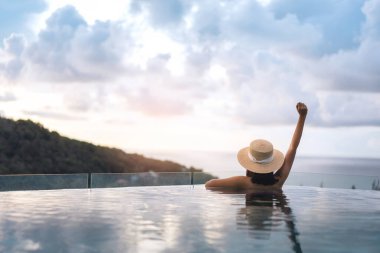 Tatil konseptiyle yaz seyahati. Asyalı bir kadının arka görüntüsü. Açık kollu. Açık havuz tatil beldesi deniz manzaralı gün batımı gökyüzü. Hasır şapka ve bikiniyle. Yerli Doğa Turizmi Phuket Adası, Tayland
