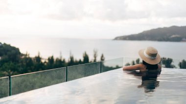 Tatil konseptiyle yaz seyahati. Asyalı kadının arka görüntüsü açık havuz tatil köyü deniz manzarasında rahatlıyor. Hasır şapka ve bikiniyle. Phuket Adası, Tayland Yerli Turizm