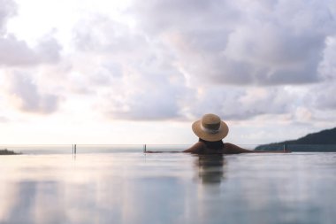 Tatil konseptiyle yaz seyahati. Asyalı kadının arka görüntüsü açık havuz tatil köyü deniz manzarasında rahatlıyor. Hasır şapka ve bikiniyle. Phuket Adası, Tayland Yerli Turizm