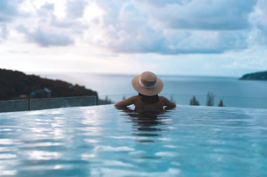 Güneydoğu tropik konseptinde yaz tatili. Arkadan bakan Asyalı kadın, açık havuz tatil köyü deniz manzarasında rahatlıyor. Hasır şapkalı ve dramatik günbatımı gökyüzüyle. Phuket Adası, Tayland