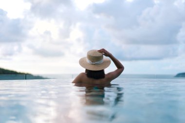 Güneydoğu tropik konseptinde yaz tatili. Arkadan bakan Asyalı kadın, açık havuz tatil köyü deniz manzarasında rahatlıyor. Hasır şapkalı ve dramatik gökyüzüyle. Phuket Adası, Tayland